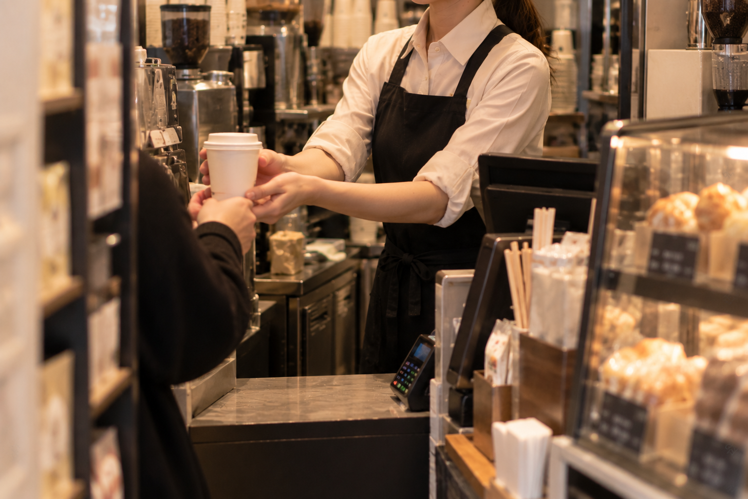 黒エプロンの女性スタッフがカウンター越しにお客さまへコーヒーカップを手渡す様子