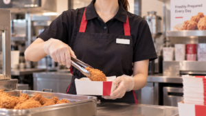 黒×赤エプロンのユニフォームで、揚げたてのオリジナルチキンをトングで持ち上げるKFCスタッフの様子