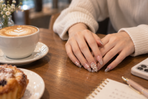 カフェのテーブルで、デコレーションネイルの手が組まれているシーン。ラテアートのカフェラテとマフィンが添えられている