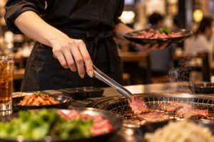 黒エプロン姿の飲食店スタッフが、上品なネイルの手でトングを持ち焼肉を焼く様子